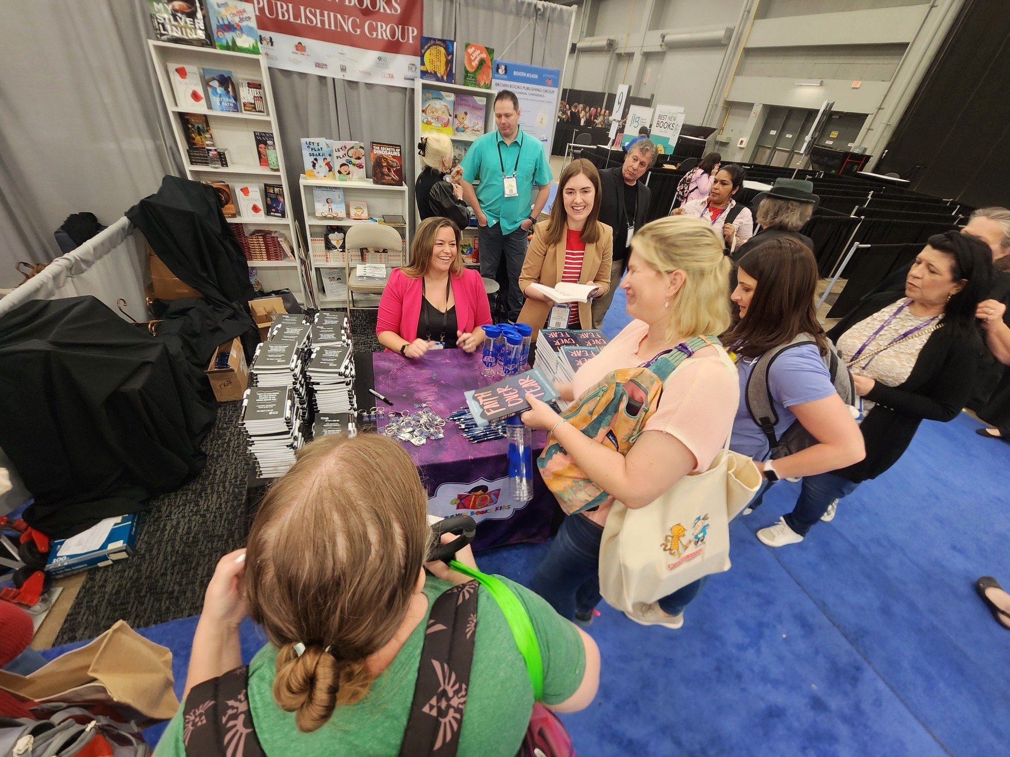 Book signing event with authors at a booth, books and merchandise on table, people waiting in line, and a woman signing books.