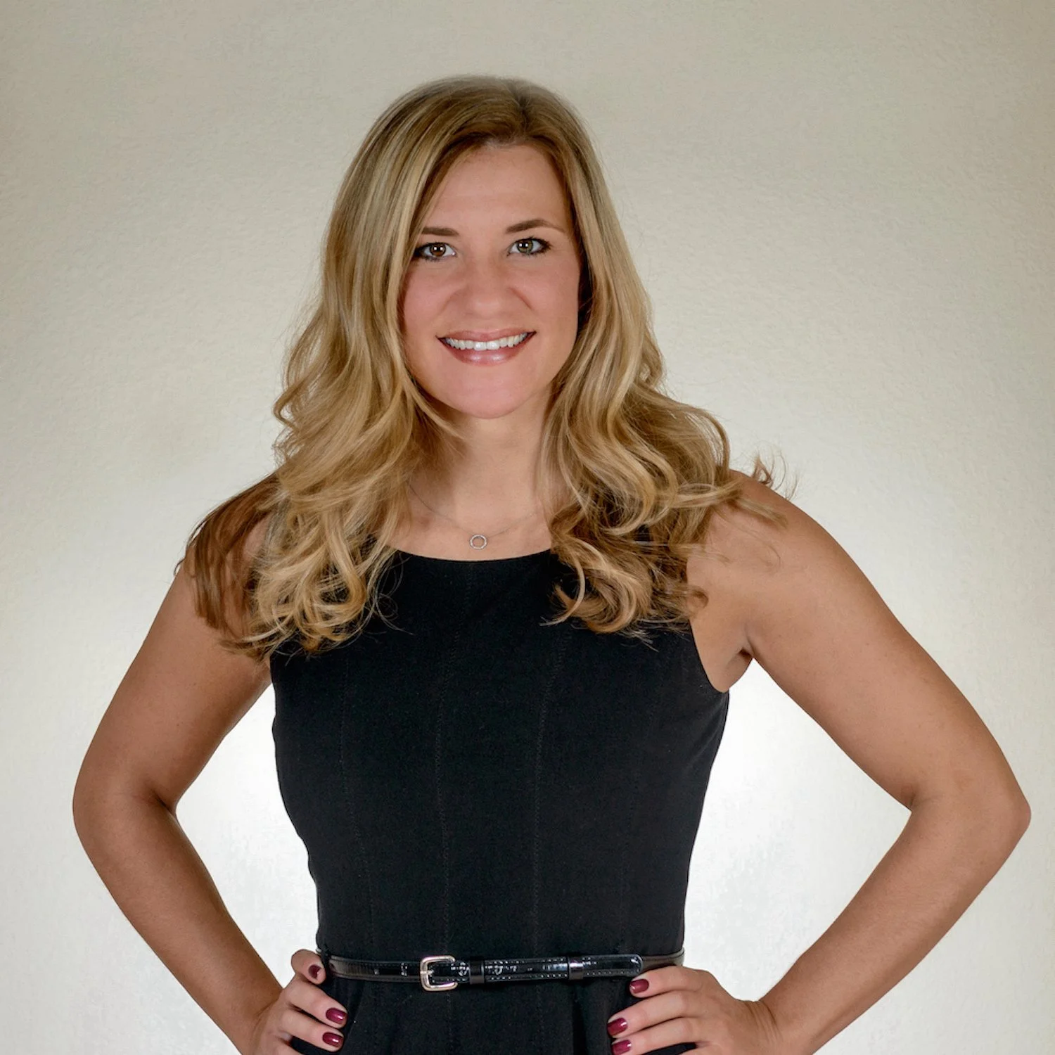 A woman with long wavy blonde hair, wearing a black sleeveless dress and a thin belt, smiling and standing against a plain light-colored background.
