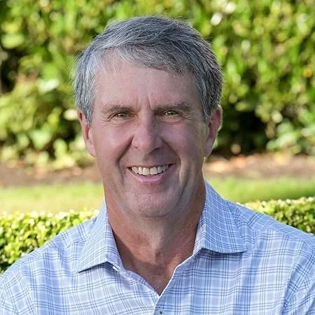 A smiling middle-aged man with gray hair wearing a light blue checkered shirt outdoors with green bushes in the background.