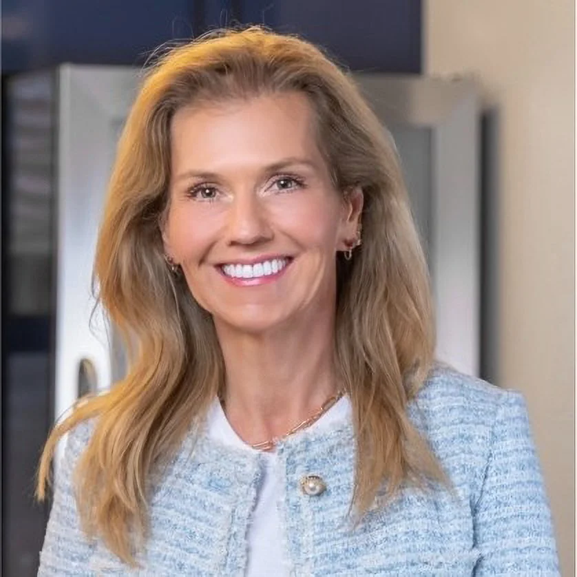 Portrait of a smiling woman with blonde hair, wearing a light blue and gray blazer and jewelry, indoors.