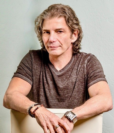 A middle-aged man with wavy gray hair and fair skin, wearing a dark gray t-shirt, sitting at a table with a light green wall in the background. He is looking at the camera with a slight smile, wearing a wristwatch and bracelets on his left wrist.