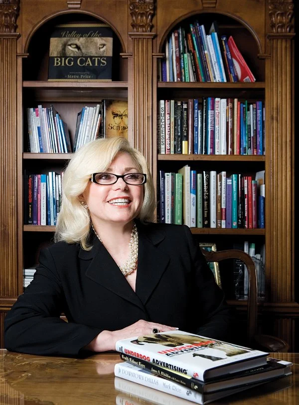 A smiling woman with blonde hair and glasses sitting at a wooden desk with a stack of books, in front of a large wooden bookshelf filled with colorful books.
