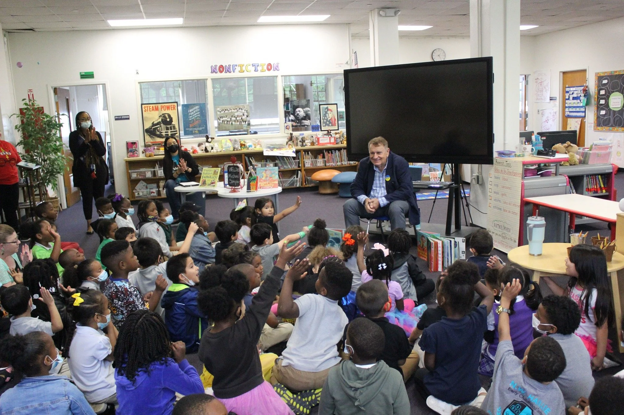 A group of young students sitting on the floor in a classroom, raising their hands while a man is seated in front of them and speaking. Several teachers and staff members are standing and sitting in the background.