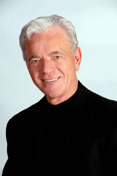 A smiling older man with gray hair, wearing a black turtleneck and blazer, against a light blue background.