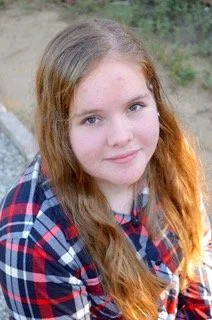 A young girl with long red hair and freckles, wearing a plaid shirt, smiling outdoors.