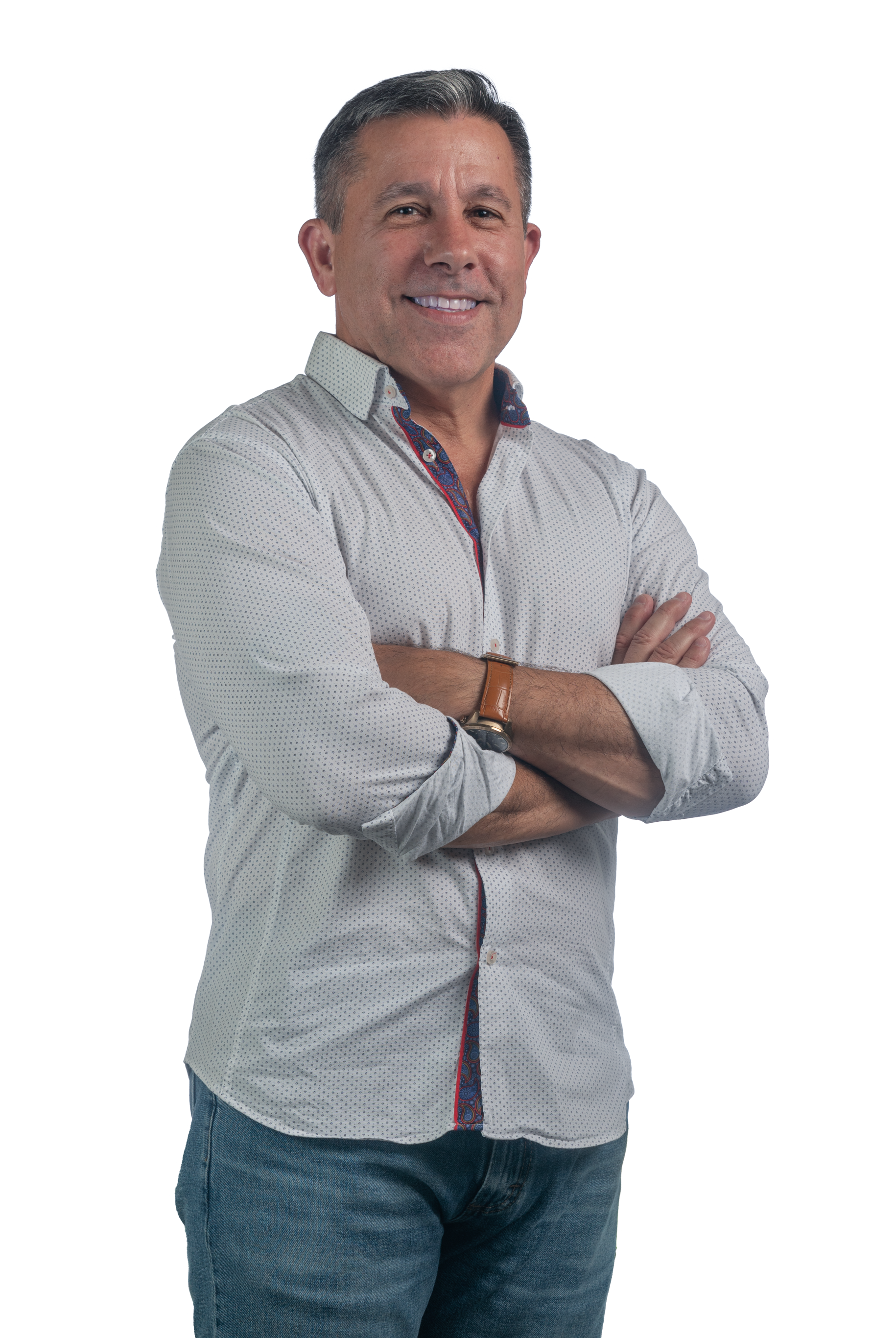 A man smiling with arms crossed, wearing a light patterned button-up shirt and jeans, against a white background.