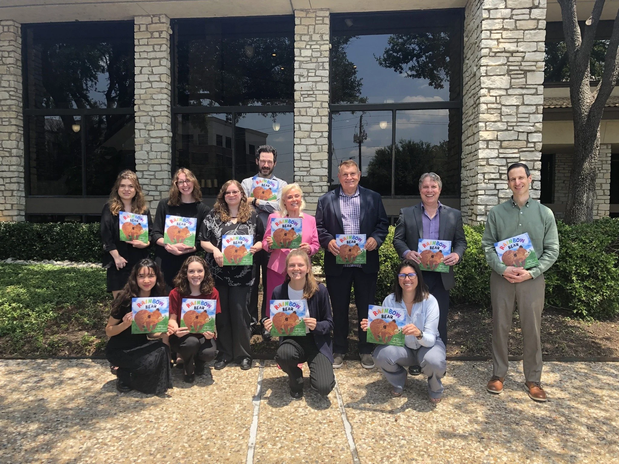 Group photo of twelve people, including children, holding children's books titled "Rainbow Bear," standing outside in front of a stone building with large windows and greenery.