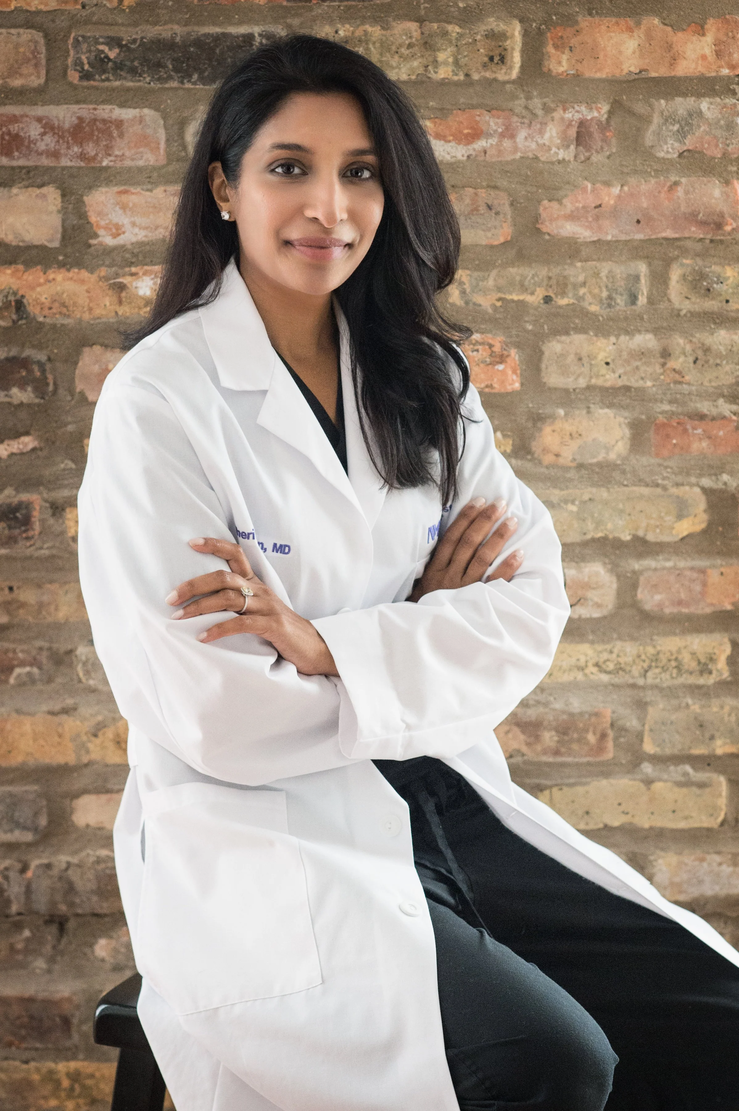 A woman in a white medical coat sitting on a stool in front of a brick wall, looking at the camera with arms crossed.