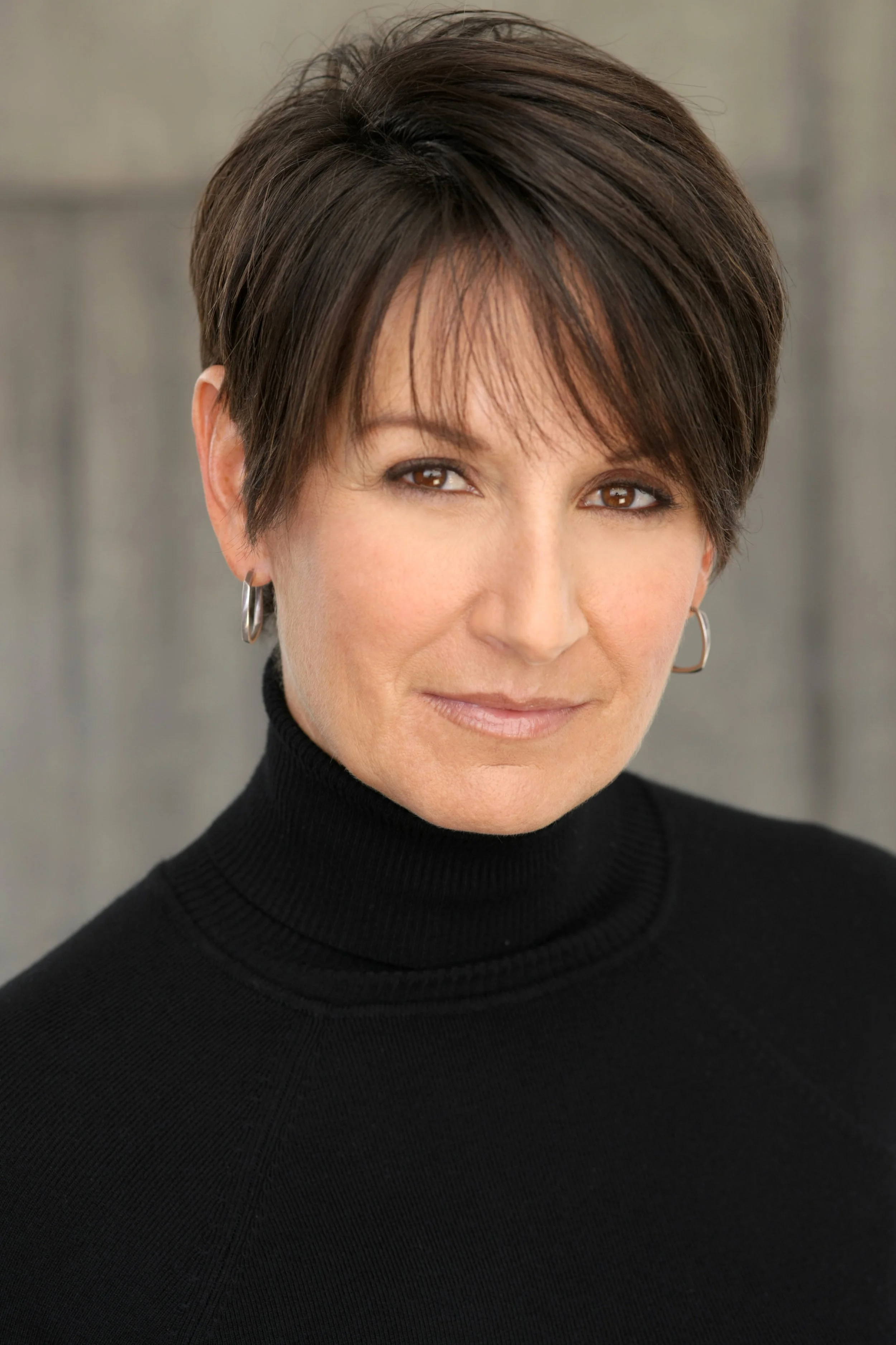Portrait of a woman with short brown hair, wearing a black turtleneck, silver hoop earrings, and smirking slightly against a plain background.