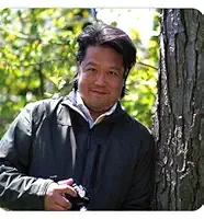 Man standing outdoors next to a tree, holding a camera, surrounded by green foliage.