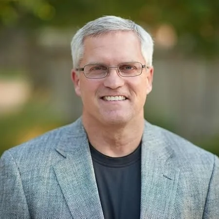 A smiling middle-aged man with gray hair and glasses, wearing a gray blazer and black shirt, standing outdoors with a blurred background of trees.