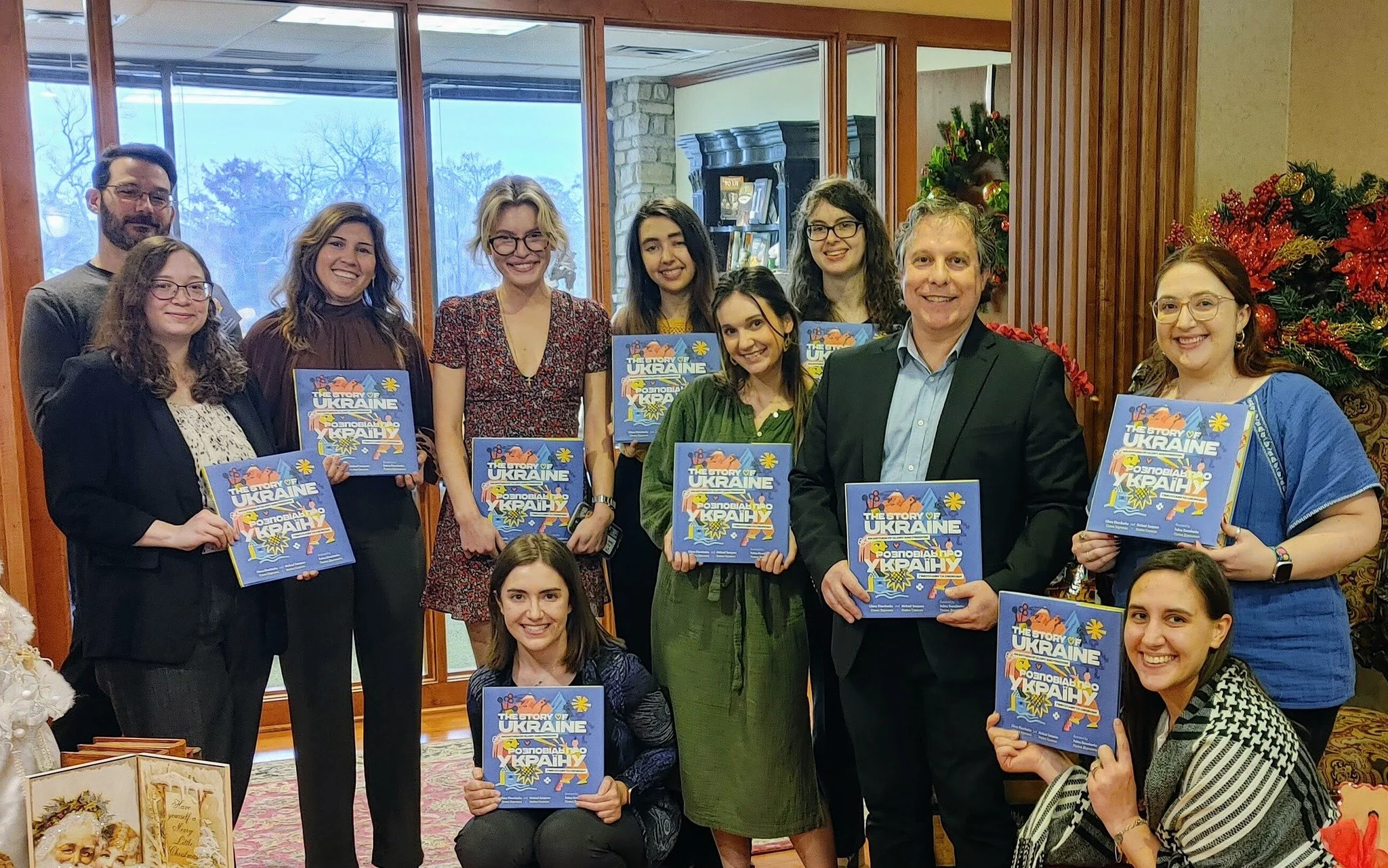 A group of eleven people, including men and women, smiling and holding books titled "The Story of Ukraine," in a cozy, decorated indoor setting with Christmas decorations.