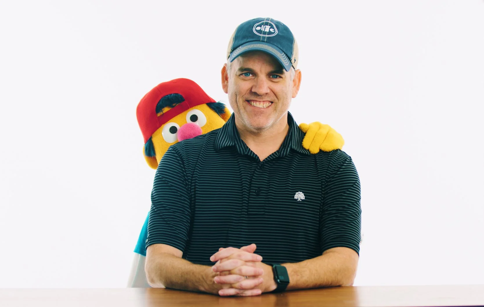 A man smiling at a table with a puppet like character standing behind him. The puppet has a yellow face, large eyes, pink nose, black eyebrows, and black hair, wearing a red baseball cap turned backward. The man is wearing a striped black polo shirt, a cap, and a smartwatch.