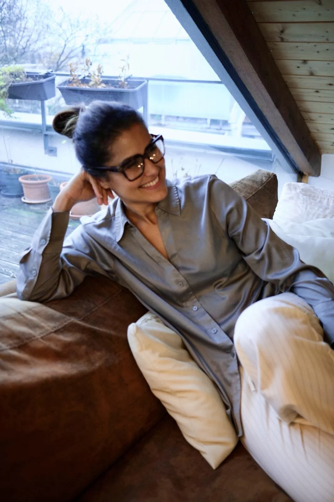 Eine lachende Frau mit Brille, die auf einer Couch in einem Raum mit Dachschräge sitzt.