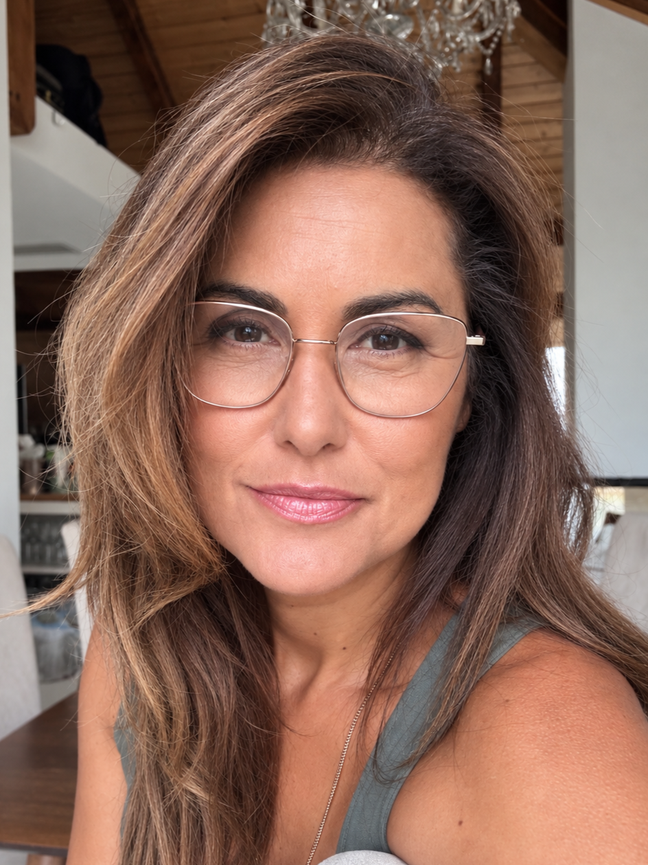 Nahaufnahme einer lächelnden Frau mit Brille und braunen Haaren, im Hintergrund ein Kronleuchter und Holzdecke.