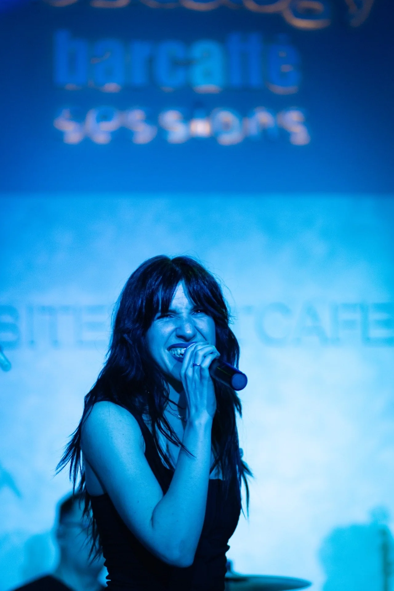 A woman with long dark hair singing into a microphone on stage, with a blue background that has some text that is blurred and unreadable.