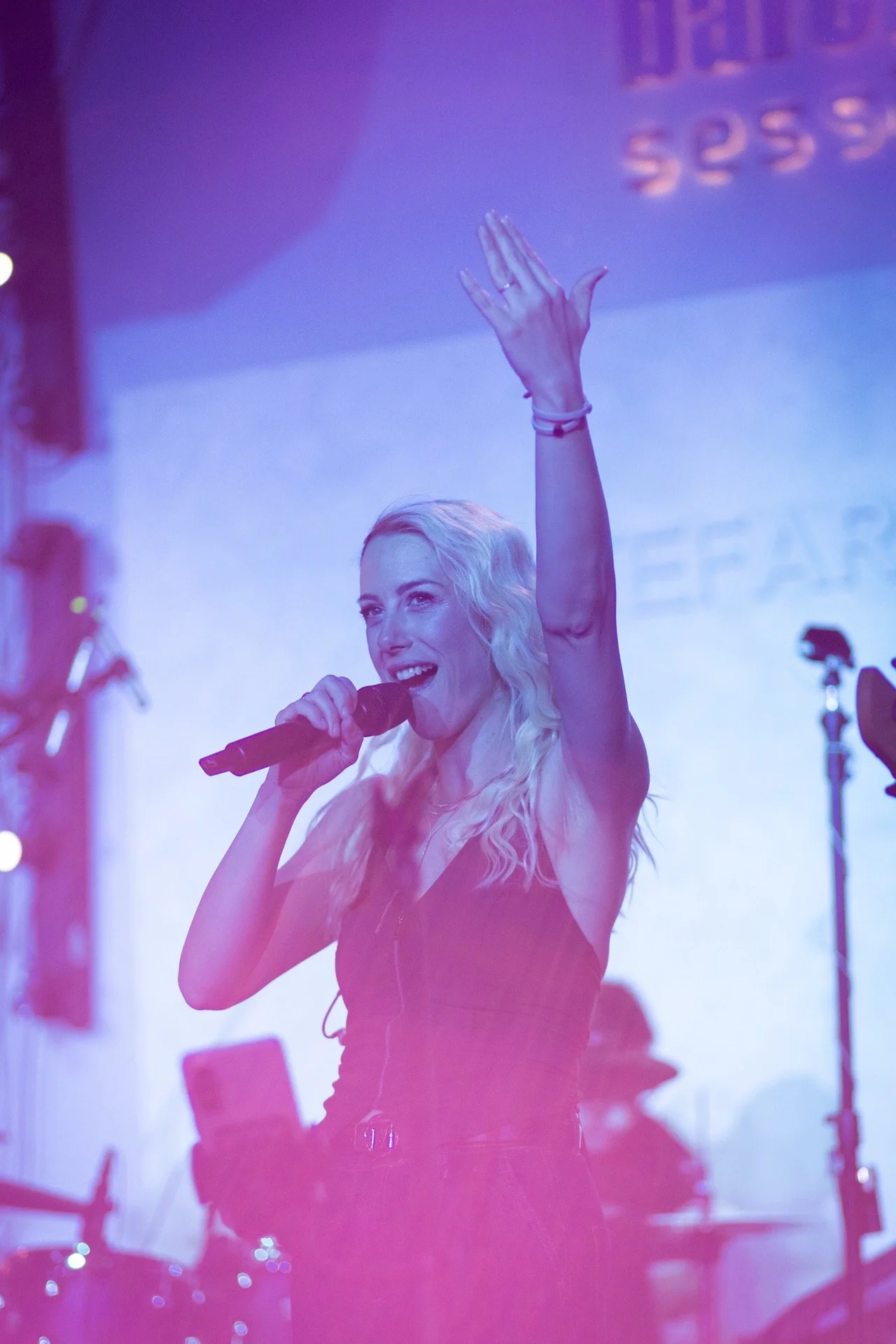 A woman with long blonde hair holding a microphone and singing or speaking into it, with her left hand raised in the air, in a dimly lit stage setting with blue and pink lighting.