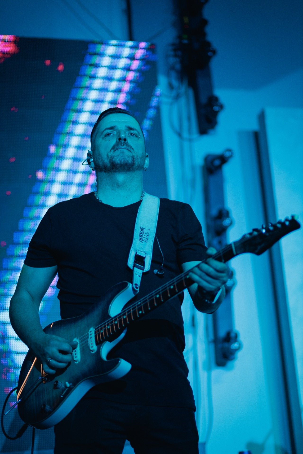 A man playing an electric guitar on stage, with blue lighting and a colorful LED backdrop.