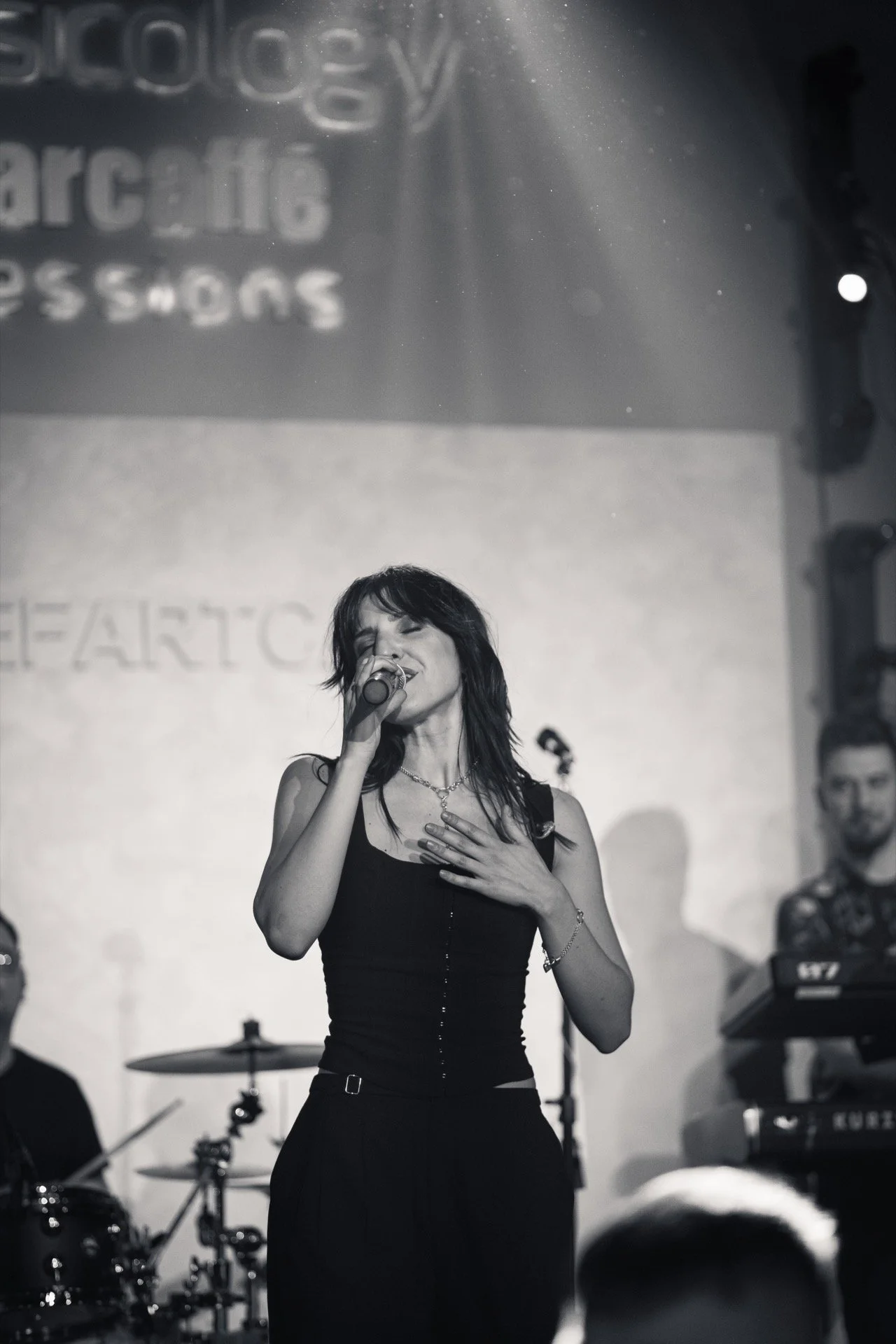 A woman singing into a microphone on stage, with a band playing in the background, during a live music performance.