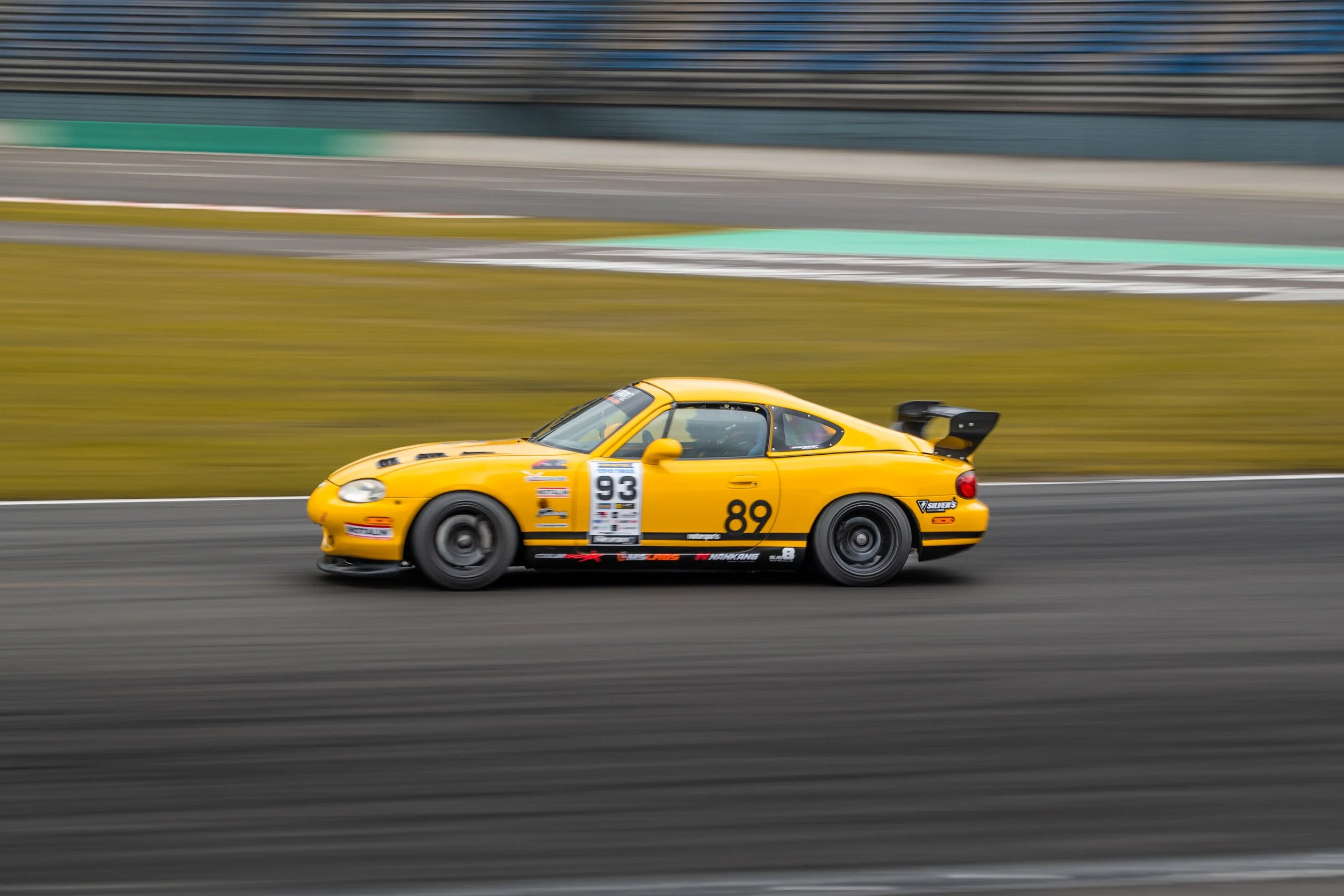 Gelber Rennwagen fährt auf einer Rennstrecke, im Hintergrund sind Tribünen und wechselnde Farbmustern zu sehen.