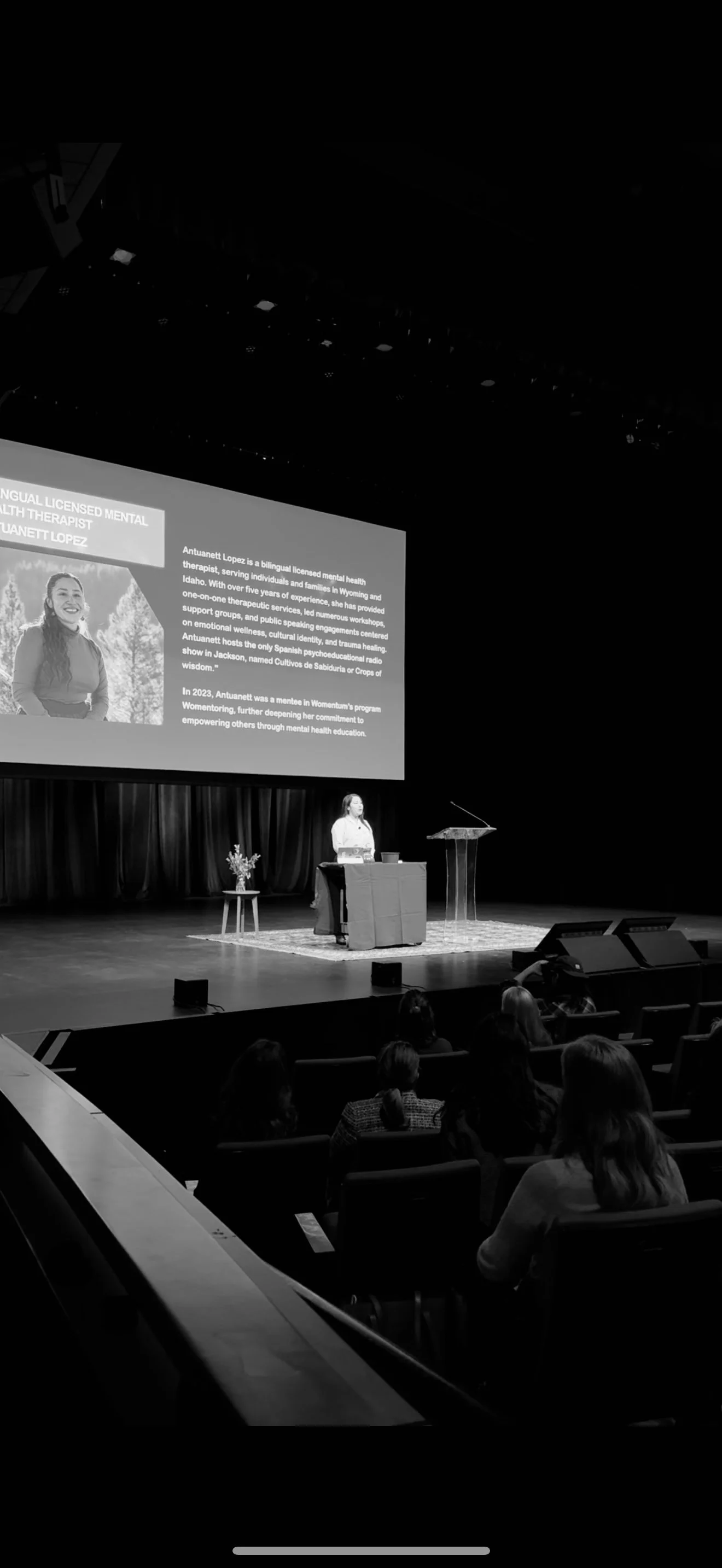 A woman giving a presentation on a stage in a theater or auditorium, with a large screen behind her displaying her biography, and an audience seated watching. The stage has a table with a flower arrangement and a transparent lectern.