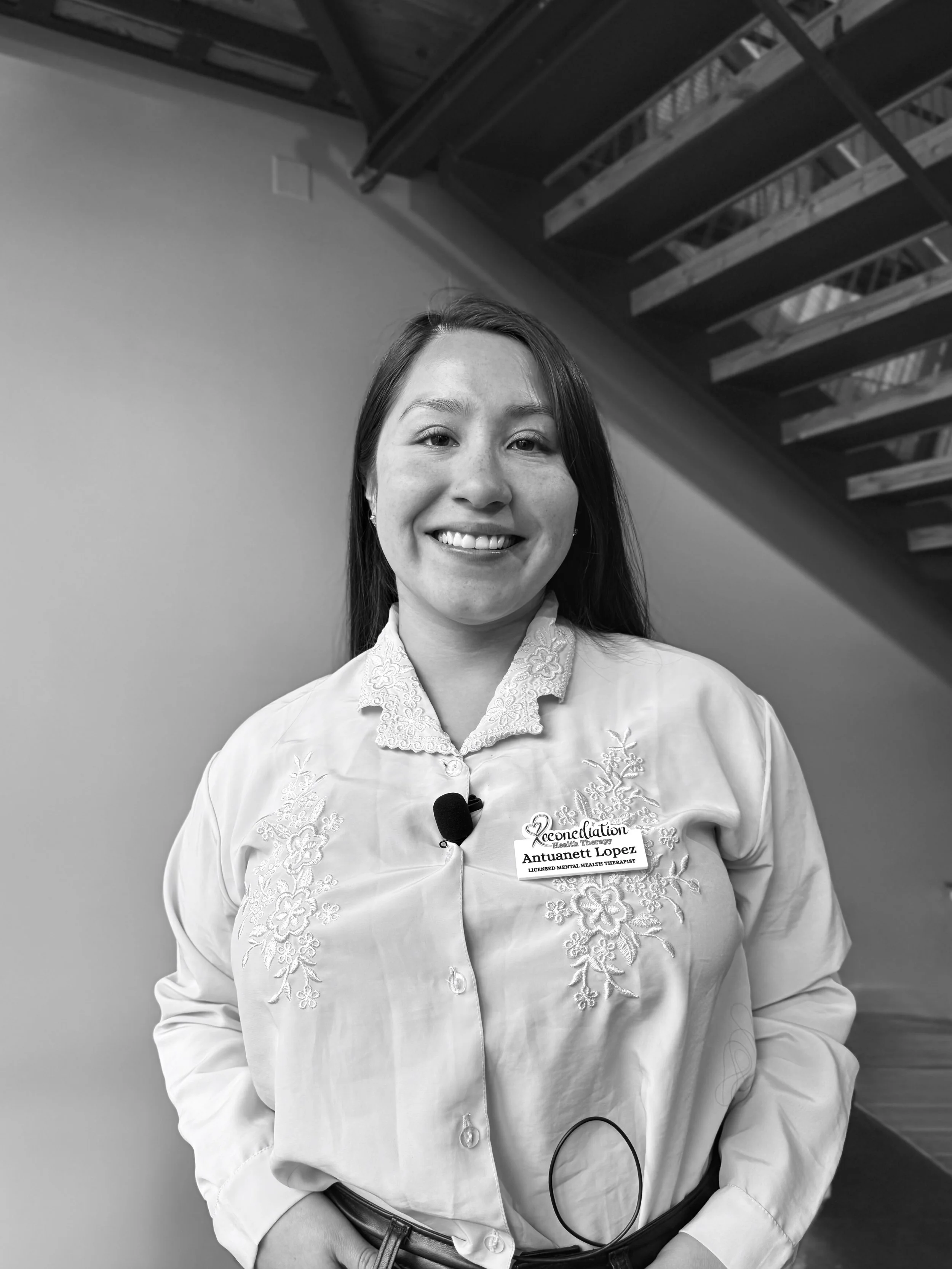 Black and white photo of a young woman smiling, wearing a embroidered blouse with a name badge, standing indoors near a staircase.