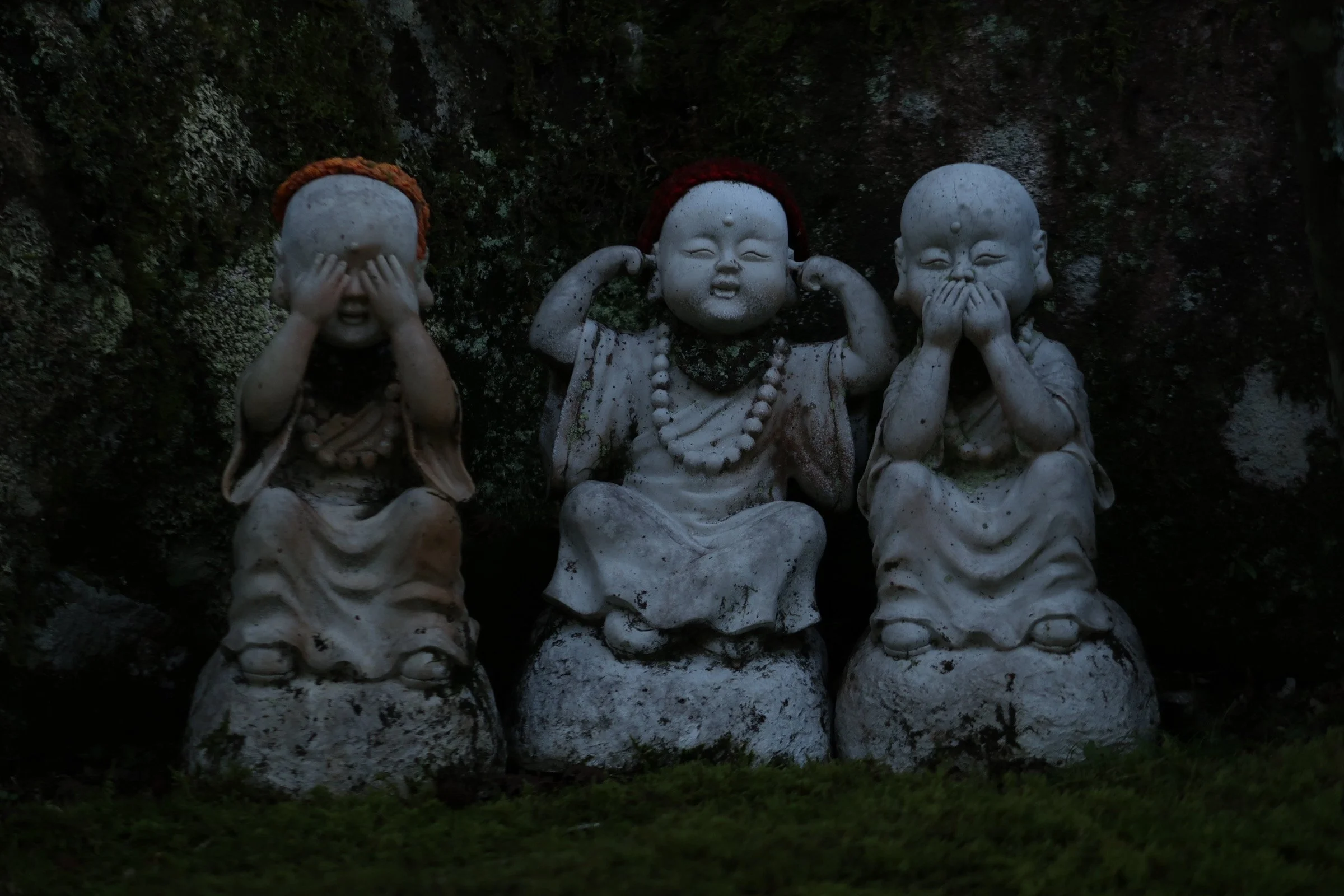 Three small stone statues of children, each covering different parts of their face or mouth, with a dark background and some greenery at the base.