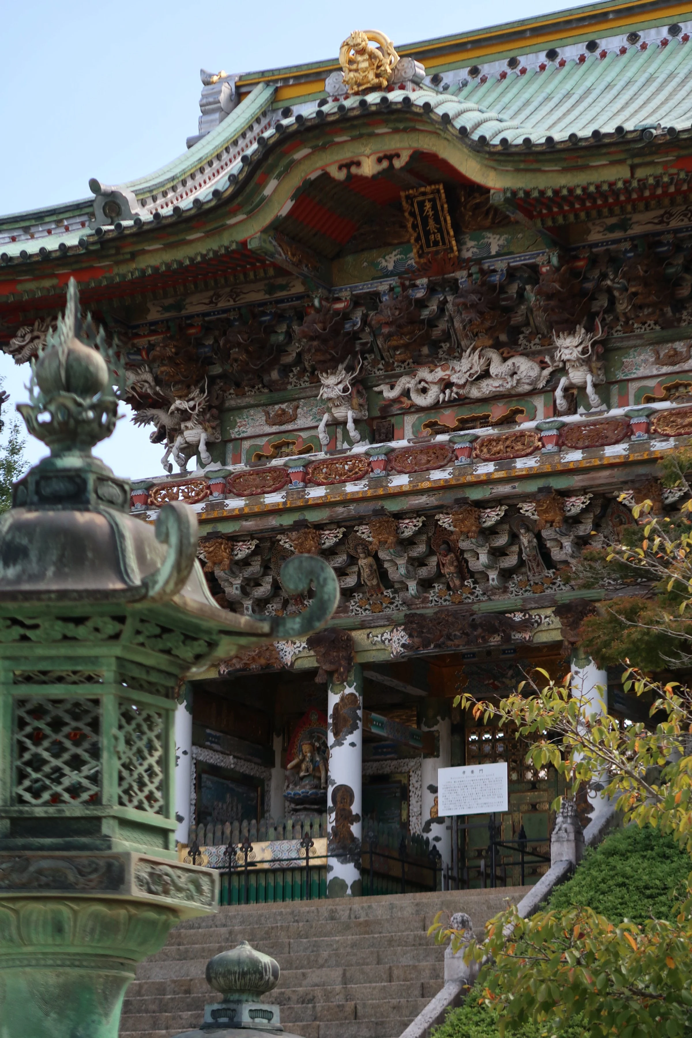 Traditional Japanese temple with ornate roof carvings, steps leading up to the entrance, and greenery, featuring a large decorative lantern in the foreground.