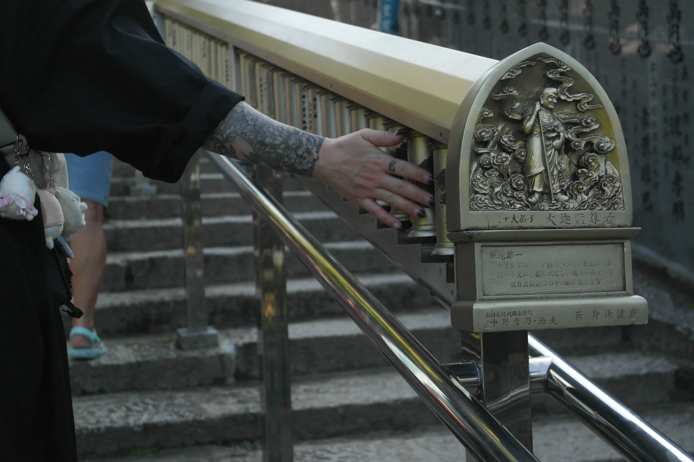 Person with tattoos on arm touching a brass railing with carved decorative plaques, positioned on stone steps, with other people in the background.