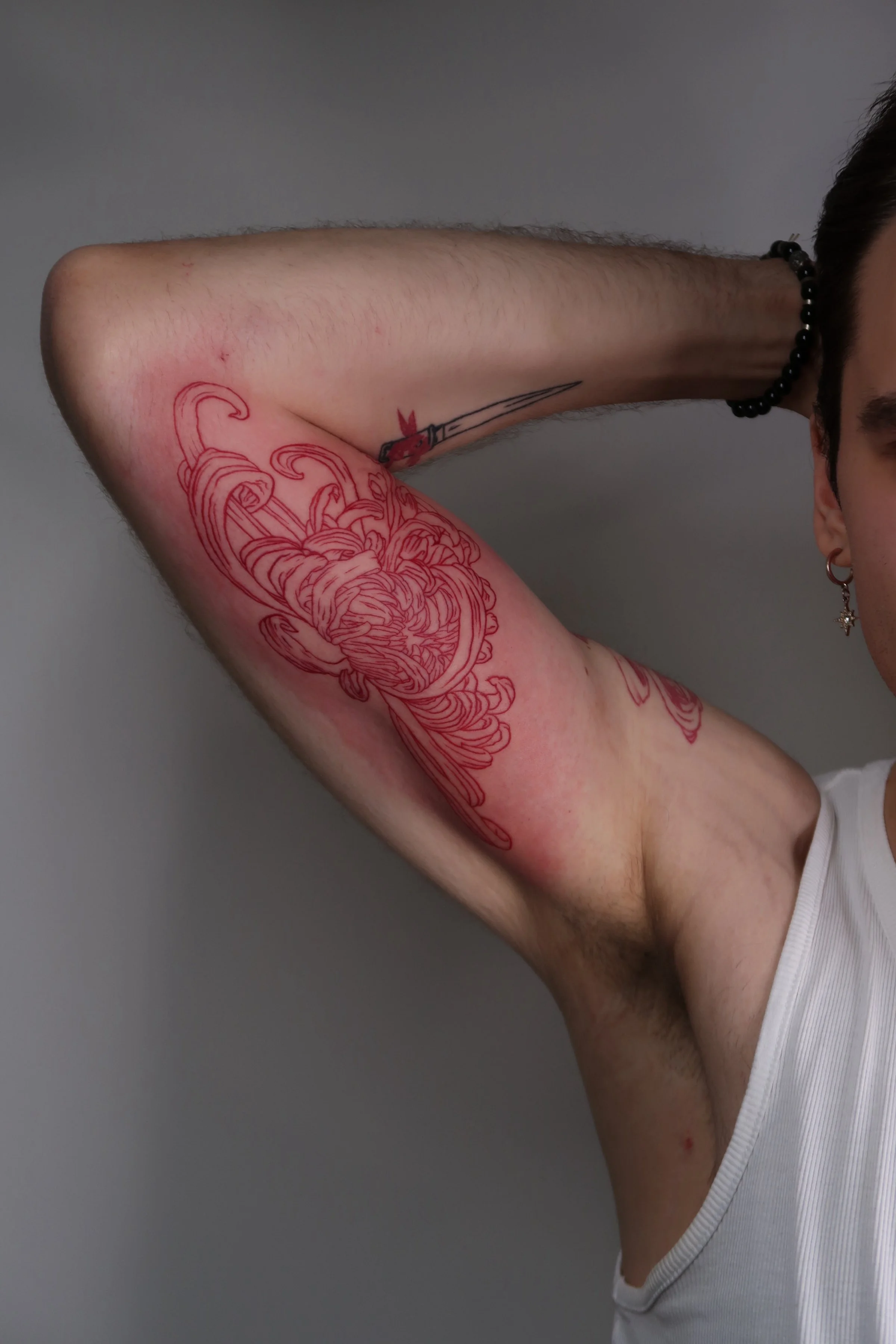 Close-up of a person's right arm with a floral tattoo outline, raised above the head, and a small sword tattoo on the inner arm. The person is wearing a white tank top, a black beaded bracelet on the wrist, and an earring.