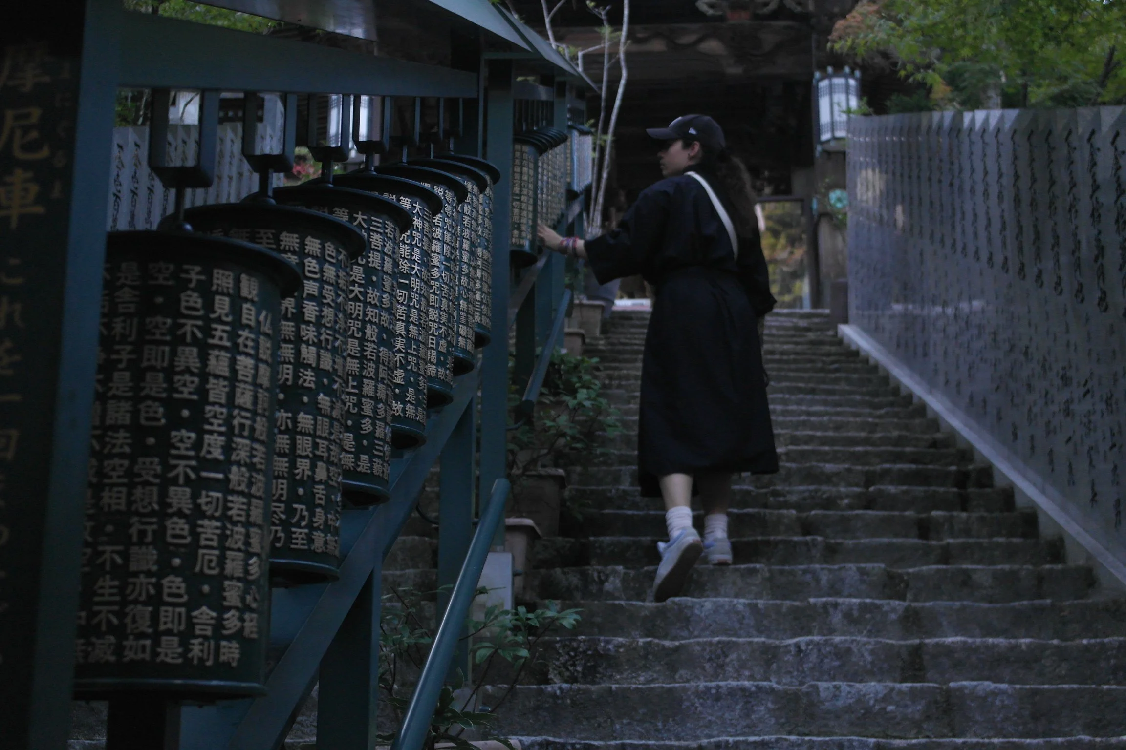 A woman in a black coat and cap ascending stone steps next to a fence with black plaques inscribed with Chinese characters on a large structure