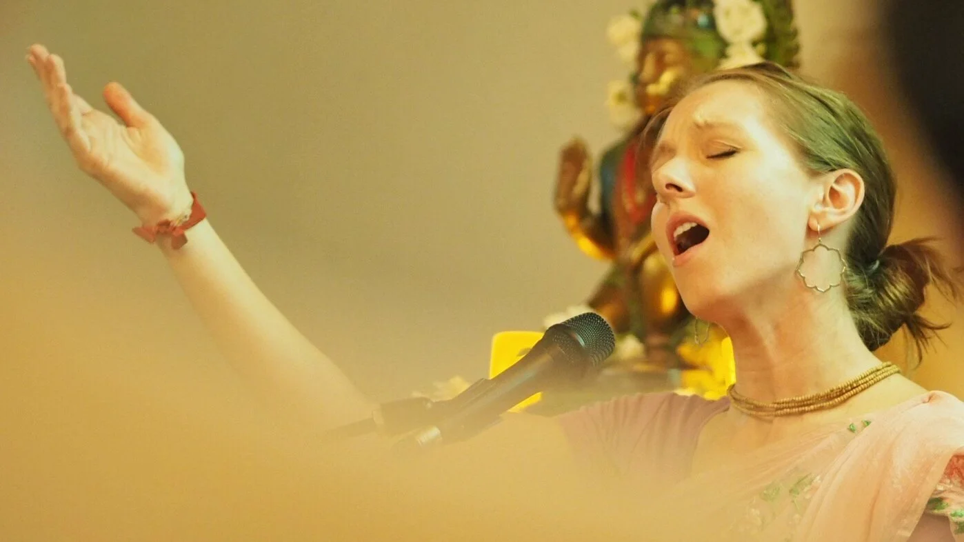 A woman singing with eyes closed, wearing traditional Indian attire with jewelry, in front of a microphone, with a decorated idol in the background.