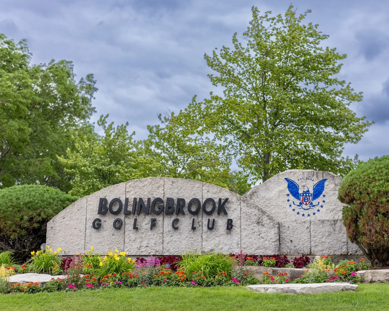 Sign for Bolingbrook Golf Club with colorful flowers in front, green bushes, and trees with green leaves, under a cloudy sky.