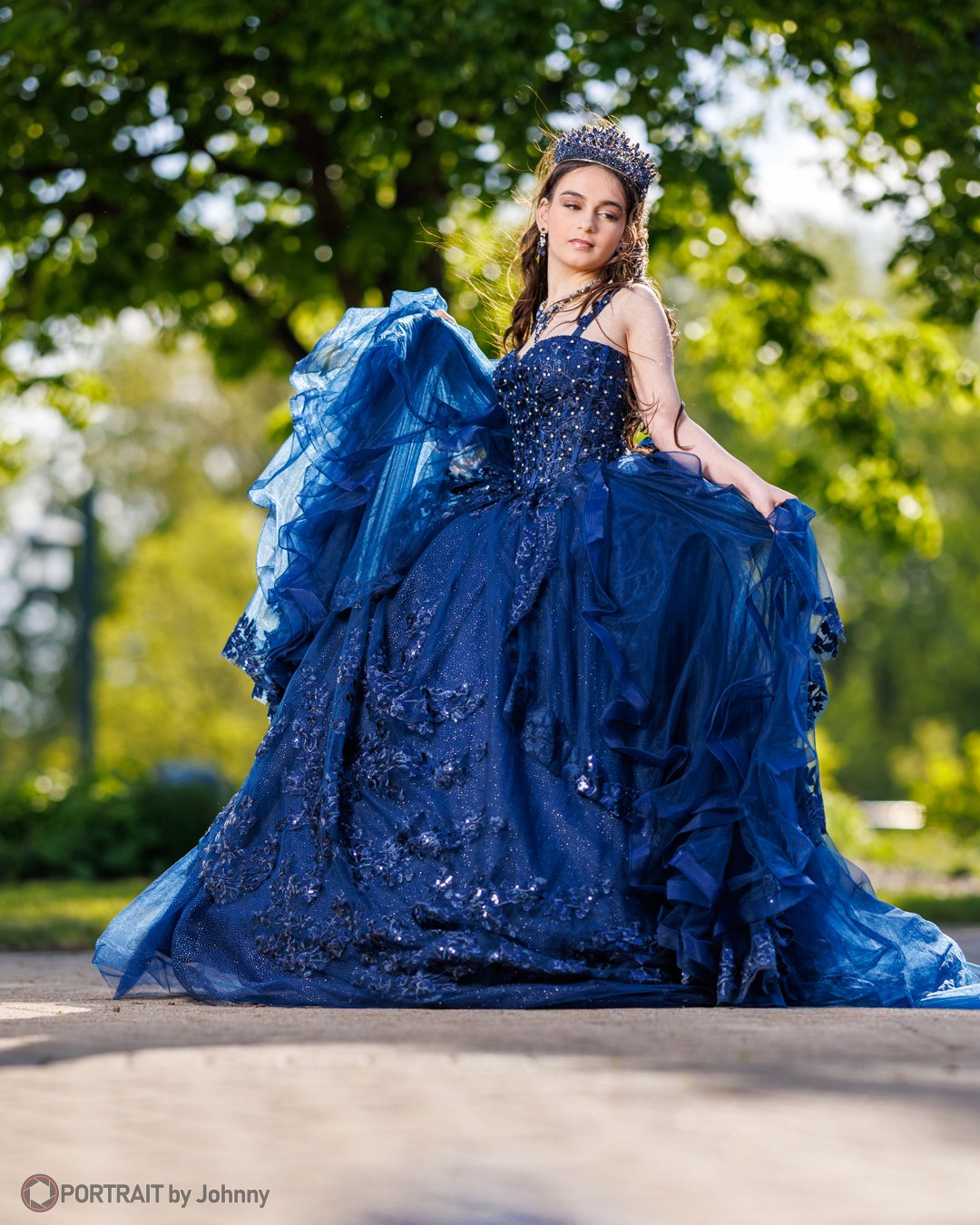 Quinceanera portrait of young woman wearing a royal blue ball gown with a tiara, standing outdoors with trees and greenery in the background.