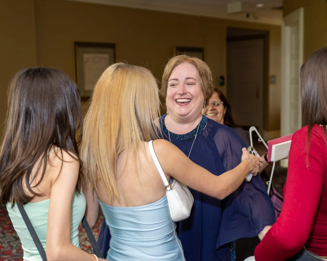 Group of women in a warm indoor setting, smiling and greeting each other, with one woman embracing another.
