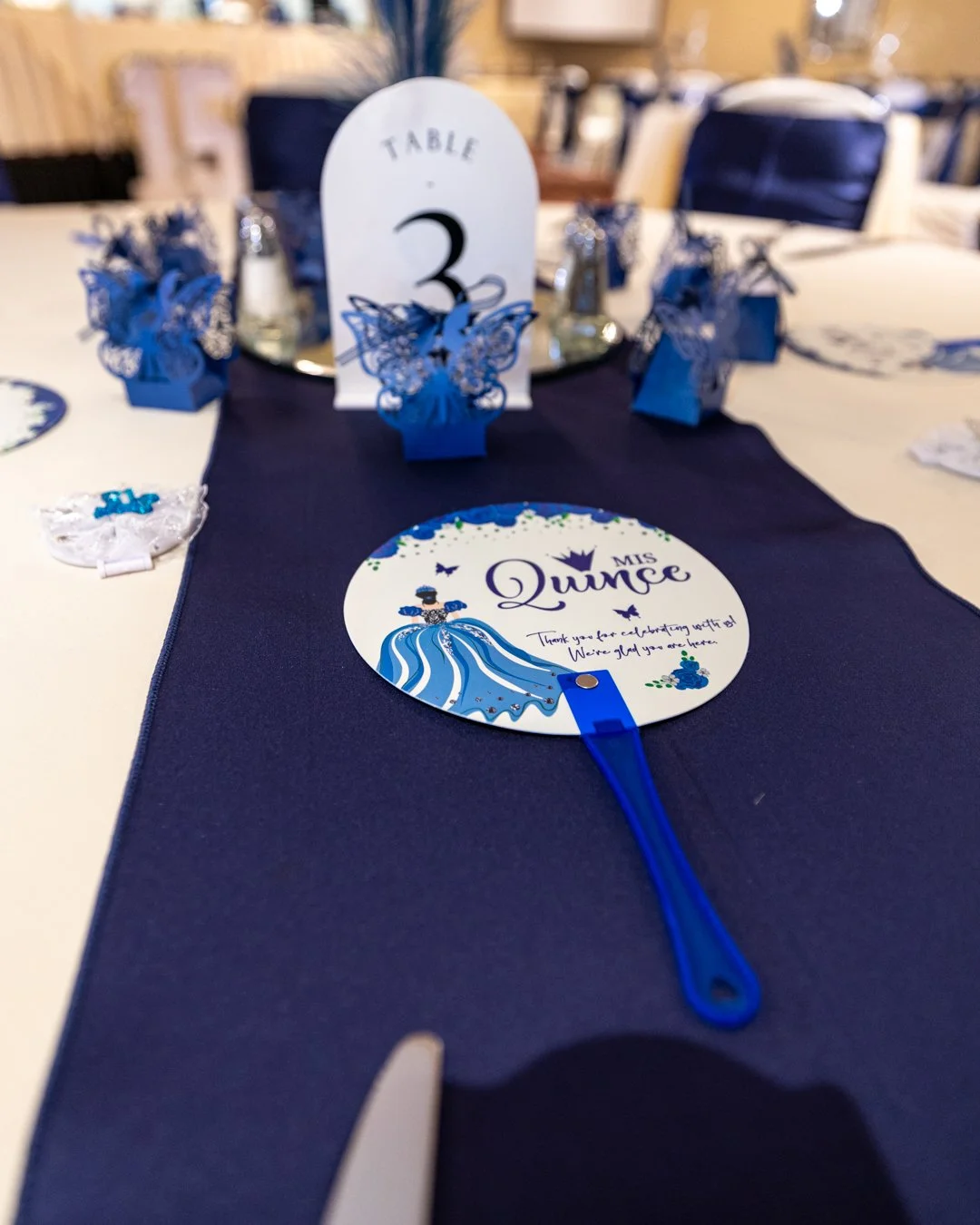 Table decorated with a blue table runner, a round sign that says 'Miss Quincee' with a princess illustration, blue gift bags with butterfly cutouts, and a table number sign indicating Table 3.
