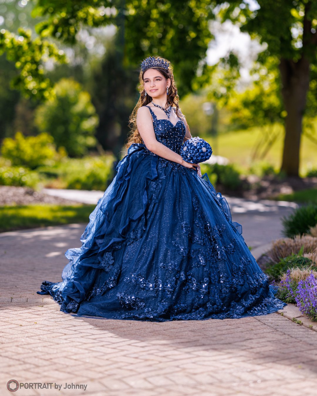 Quinceanera portrait of young girl in a dark blue, ball gown with a full skirt, accessories, and a tiara, holding a matching clutch, standing outdoors on a sunny day surrounded by greenery.