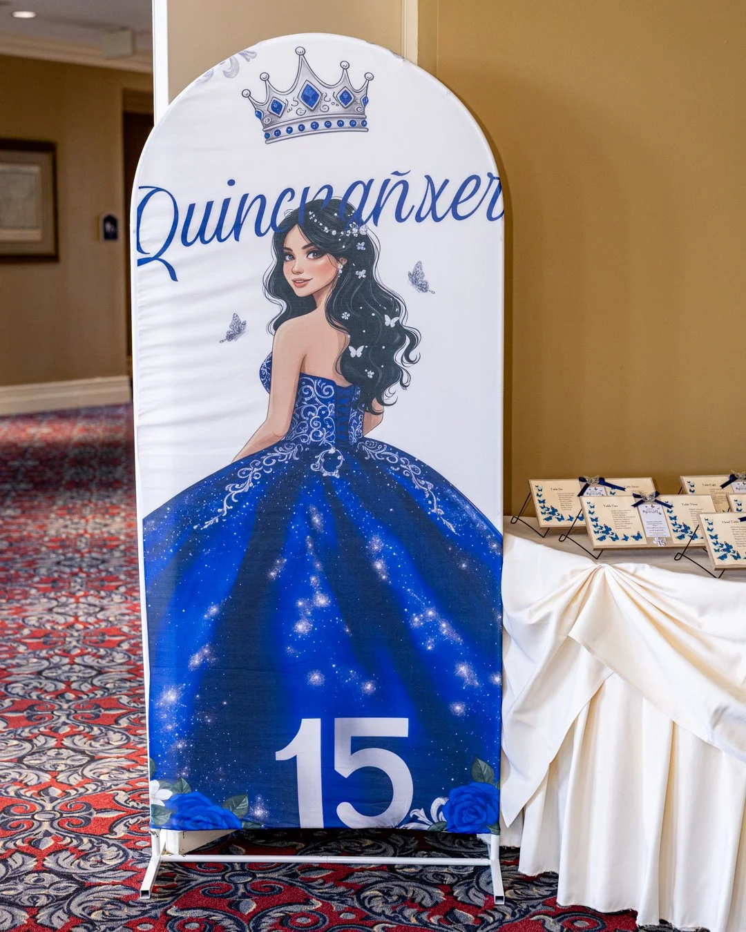 A decorative sign for a quinceañera featuring a girl in a blue, starry ball gown with long black hair, a crown, and butterflies, with the number 15 at the bottom and small tables with schedules or programs nearby.