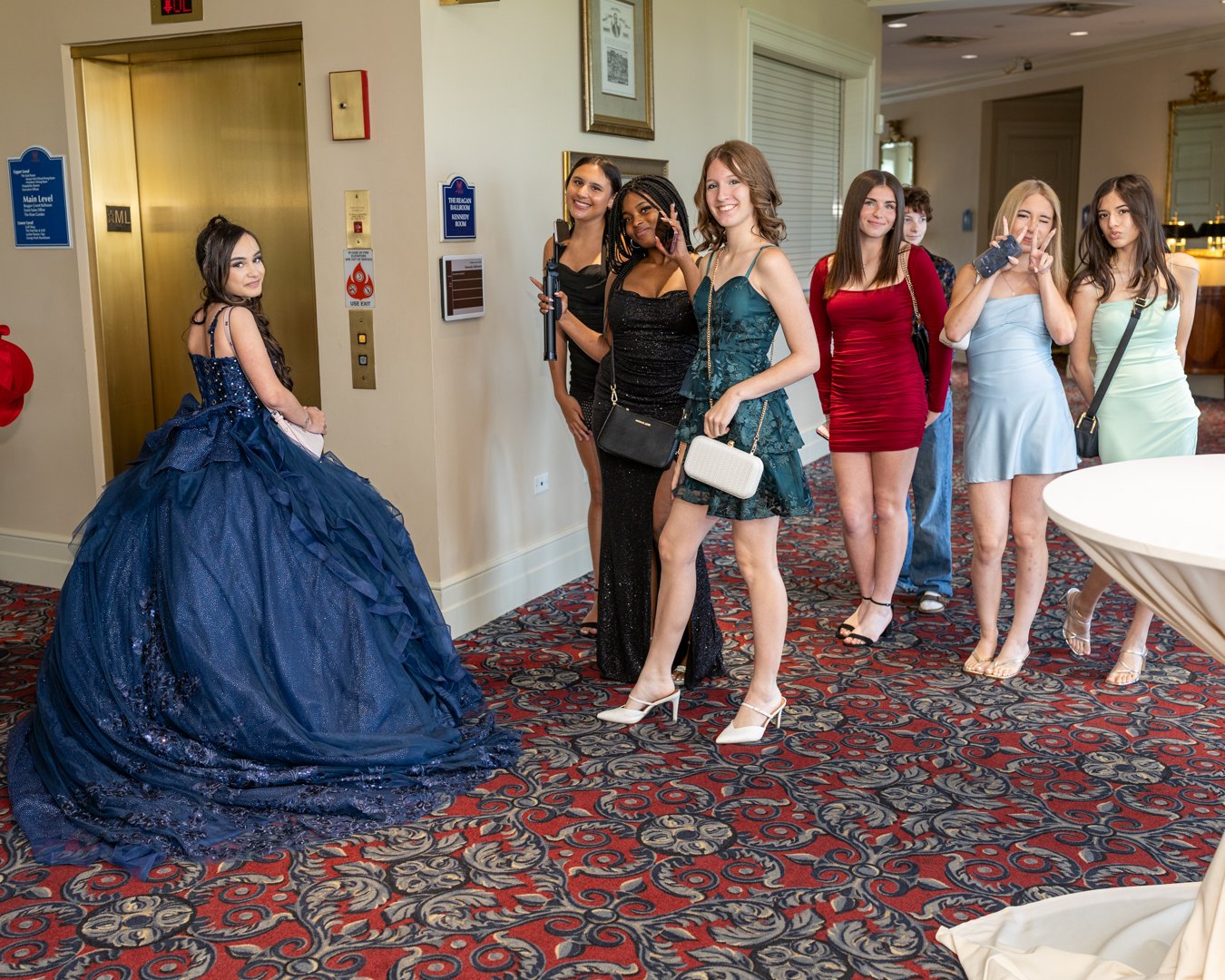 A group of seven young women dressed in formal gowns standing in a hotel lobby area, some smiling and posing, one girl in a large navy blue gown looking at the camera, while the others pose or look elsewhere.