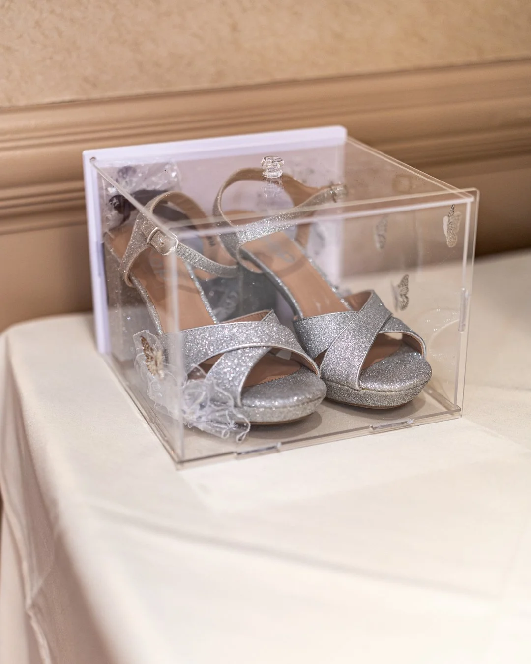 A pair of sparkly silver high-heeled shoes inside a clear plastic display box placed on a white table.