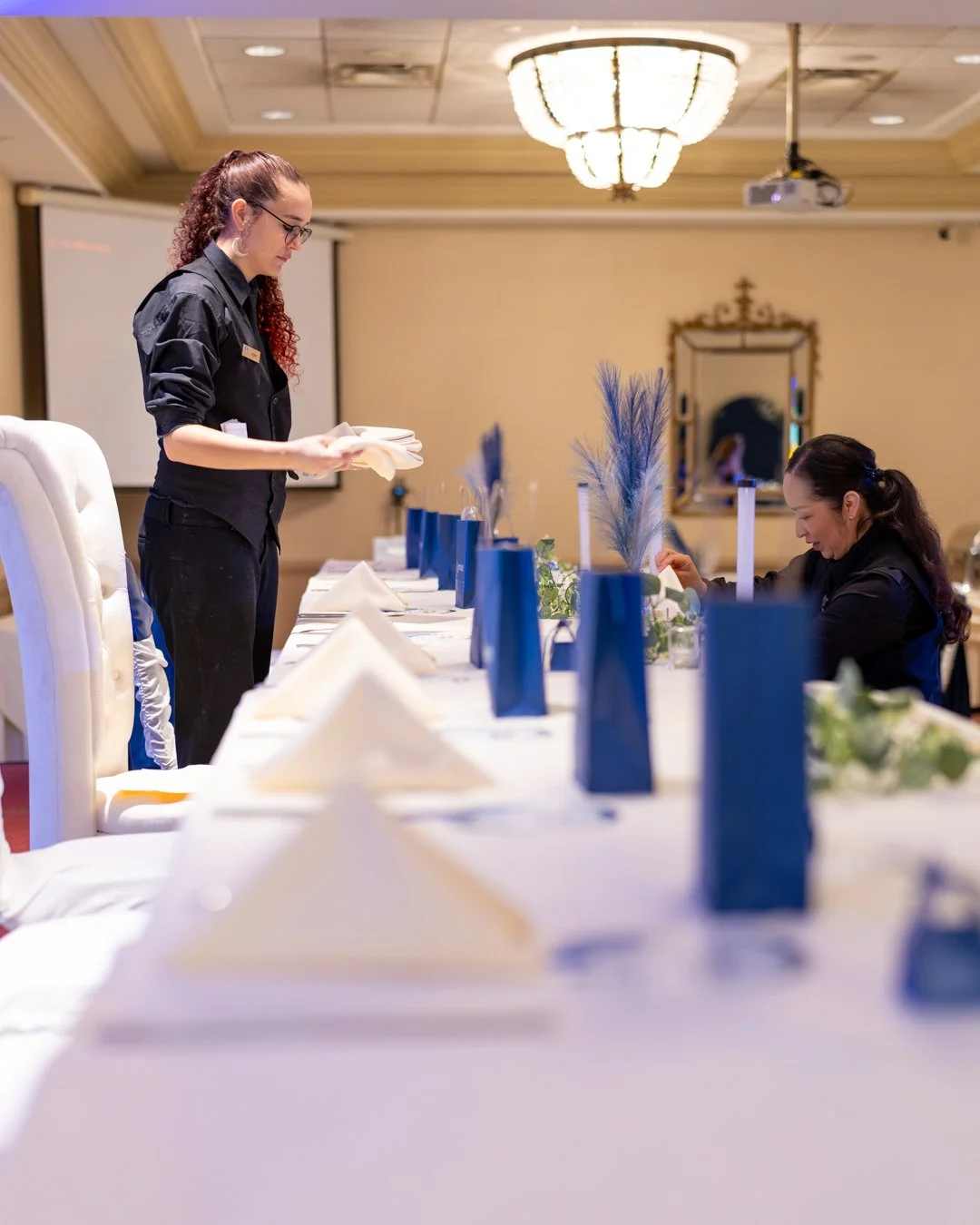 Two women preparing a formal dining table with blue gift bags, white napkins, and decorative feathers in a banquet room.