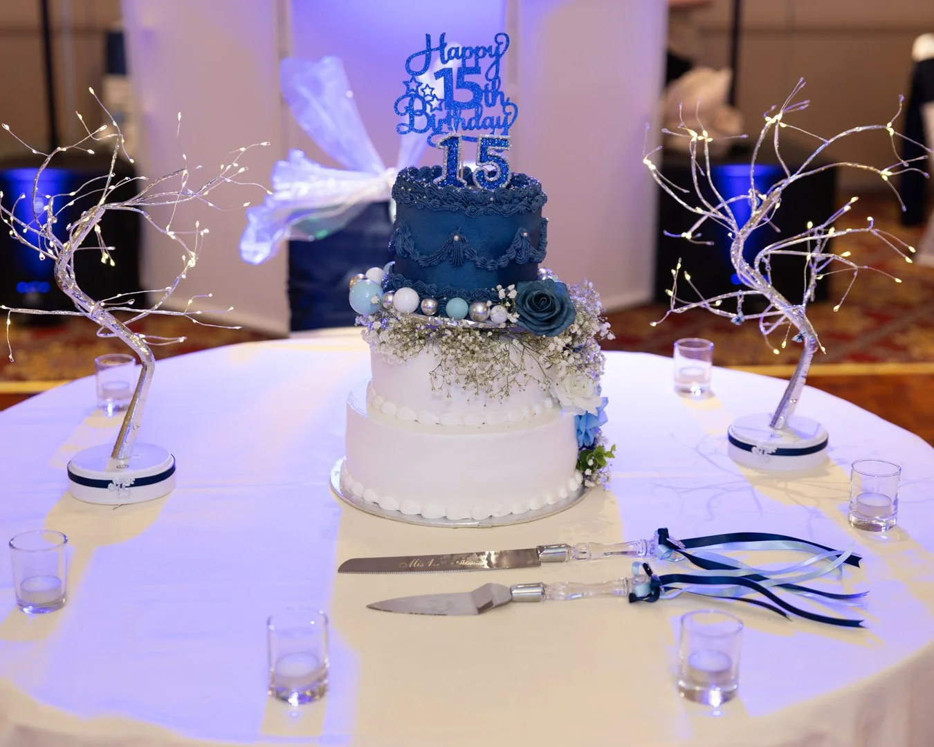 A birthday cake with blue and white frosting decorated with flowers and pearls, topped with a 'Happy 15th Birthday' topper, on a round table with small candles and lighted decorative branches.