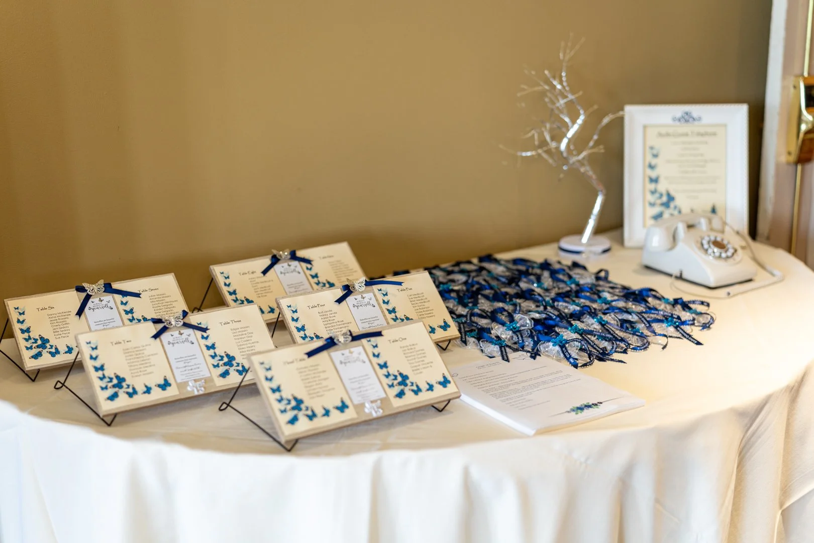 Wedding seating chart with blue butterfly accents, a decorative table with ribbons, a framed sign, a vintage rotary phone, and a decorative tree on a cream-colored tablecloth.
