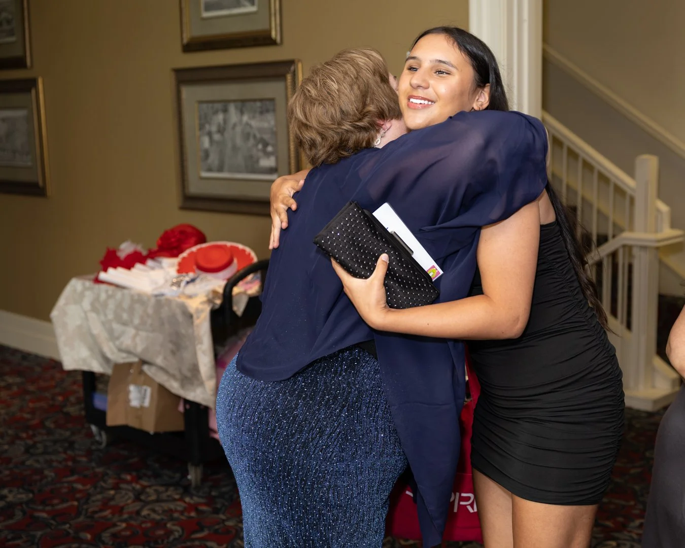 Two women hugging in an indoor setting, one wearing a black dress and holding a black clutch, the other wearing a sparkly blue dress with a brain tumor awareness ribbon, with framed photos on the wall and a table with red hats and papers in the backg