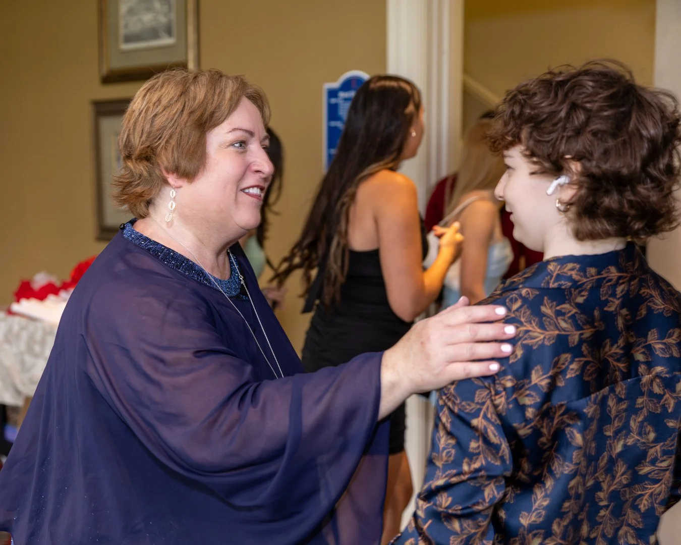 Two women are smiling and greeting each other at a social gathering indoors, with other people in the background.