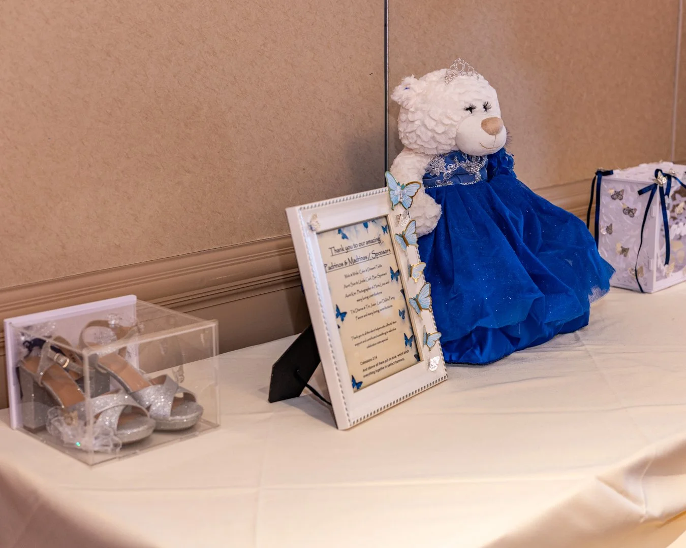 Decorative teddy bear dressed in a blue gown with a lace collar, wearing a tiara, and surrounded by thank you notes and a pair of silver glittery children's shoes on a table at a celebration or event.