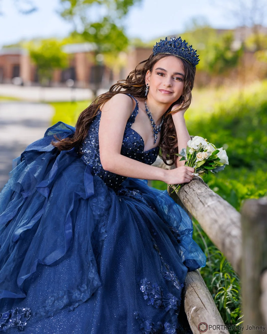 Quinceanera portrait of young woman in a blue ball gown and crown, holding a bouquet of white flowers outdoors on a sunny day.