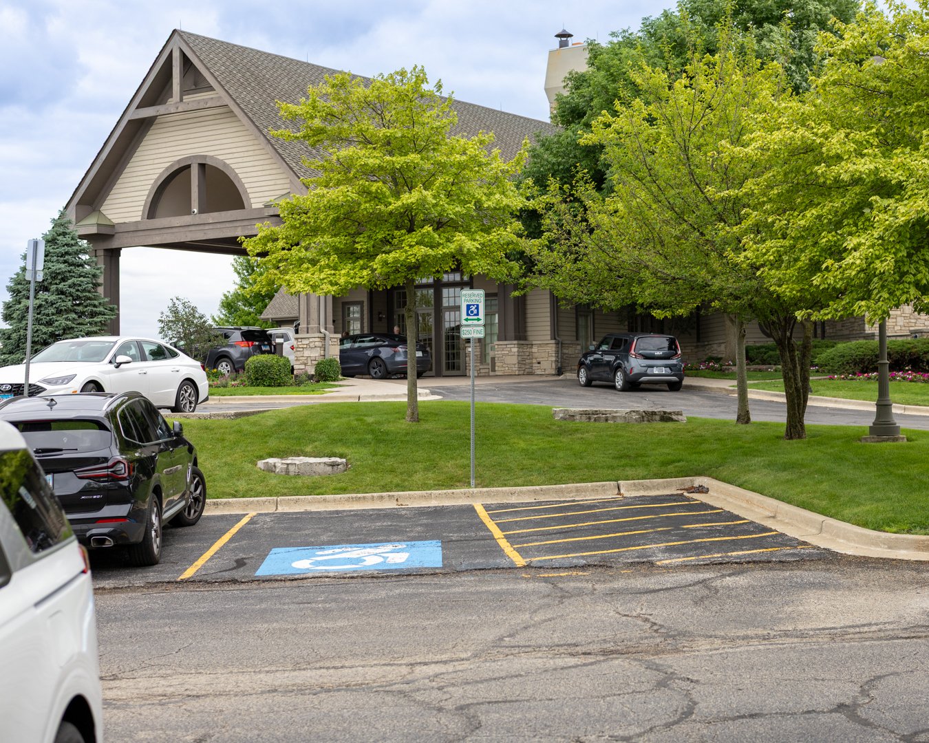 Parking lot with several cars, including one partially visible white vehicle, and one black SUV, with an accessible parking space marked with blue paint and a white wheelchair symbol. The parking lot is adjacent to a grassy area with trees and a buil