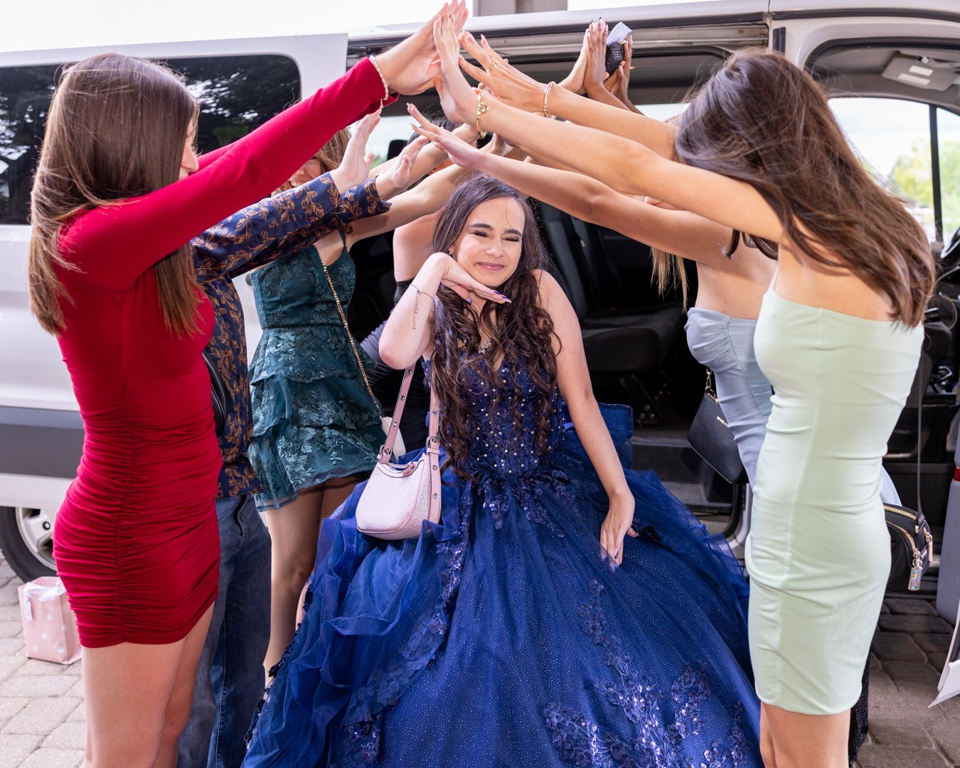 A young woman in a blue ball gown sits in front of an open van door while several women in dresses stand around her, creating an arch of hands over her head, celebrating her birthday or special occasion.