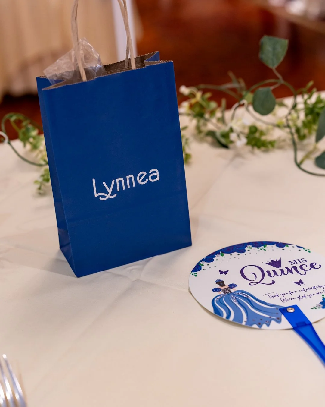 Blue gift bag with white Lynnea logo, placed on a white tablecloth. Next to it is a round card with Queen theme and the words "Ms. Queen" in purple, surrounded by butterflies and a princess illustration.